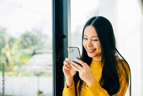 A woman near the window uses a smartphone app typing and connecting for social media. Reflecting communication online reading and texting at home.
