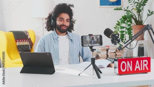 Young enthusiastic Arabian man blogger shoots video on mobile phone camera and applauds announcing results competition on page on social networks sits at table with microphone in home office. On air photo