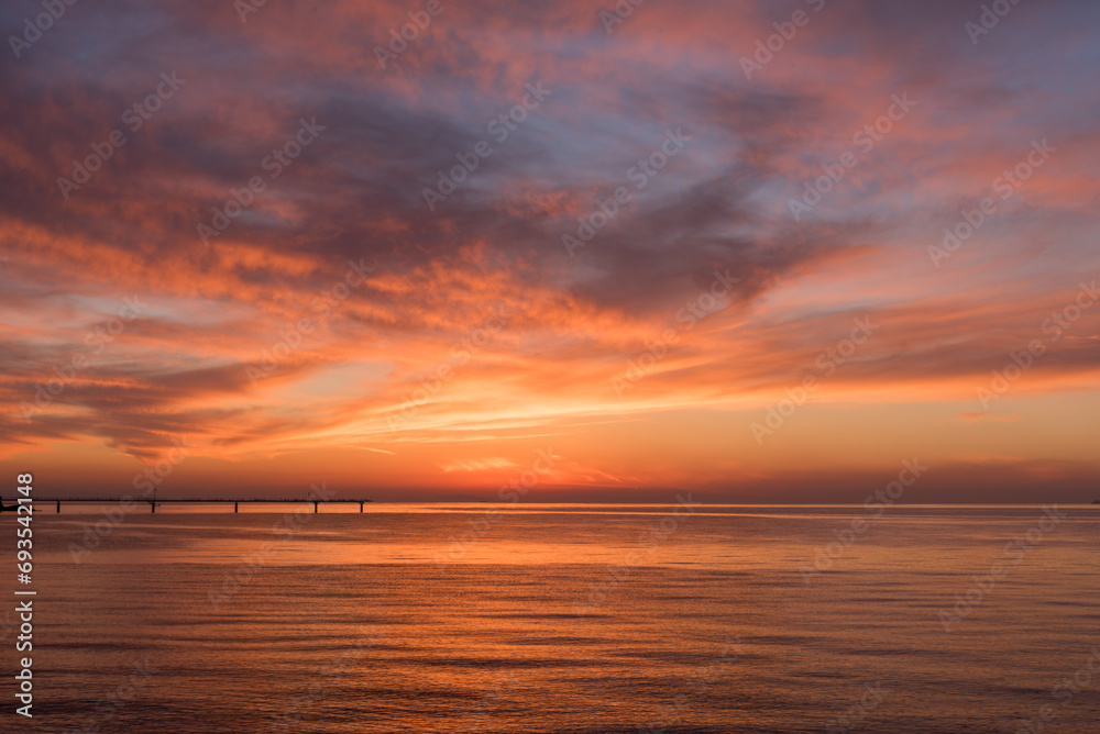 Obraz premium 遠くに桟橋の見える、赤く染まった夕暮れの海と美しい空