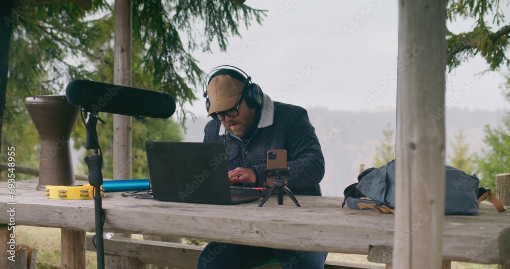 Caucasian musician in headphones uses MIDI keyboard to compose song sitting in gazebo in forest. Adult man creates and records music using phone on tripod and laptop during vacation in mountains.