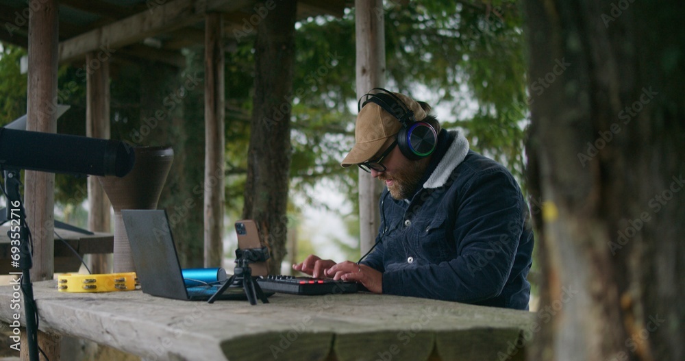 Male musician in headphones plays on MIDI keyboard, creates music and moves rhythmically sitting in gazebo in forest. Adult Caucasian man uses laptop and phone on tripod to record song outdoors. Male musician in headphones plays on MIDI keyboard, creates music and moves rhythmically sitting in gazebo in forest. Adult Caucasian man uses laptop and phone on tripod to record song outdoors.
