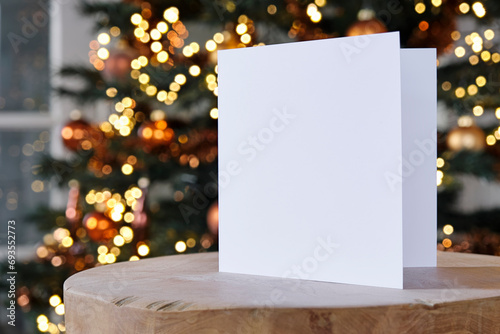 Standing blank empty square greeting card mock up before a blurred christmas tree with copy space. For use as a Christmas, background template. 