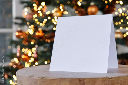 Standing blank empty square greeting card mock up before a blurred christmas tree with copy space. For use as a Christmas, background template.    