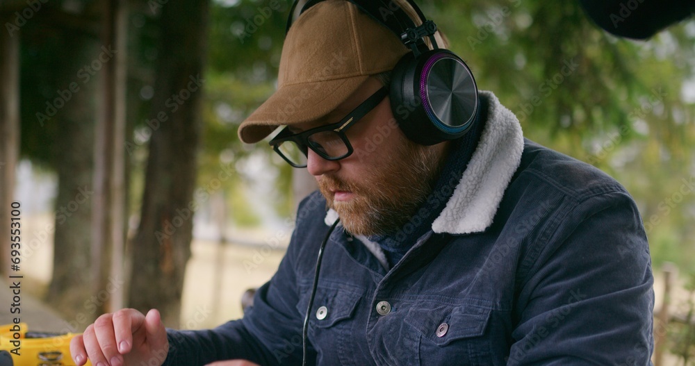 Adult male musician sits in wooden gazebo, sings and creates new composition. Traveler in glasses and headphones remotely works during vacation in beautiful forest. Music production outdoor concept. Adult male musician sits in wooden gazebo, sings and creates new composition. Traveler in glasses and headphones remotely works during vacation in beautiful forest. Music production outdoor concept.