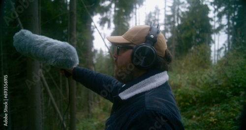 Male tourist wearing headphones records sounds of nature for movie in coniferous wood using furry windscreen microphone. Sound engineer works with professional audio equipment outdoors. Film industry.