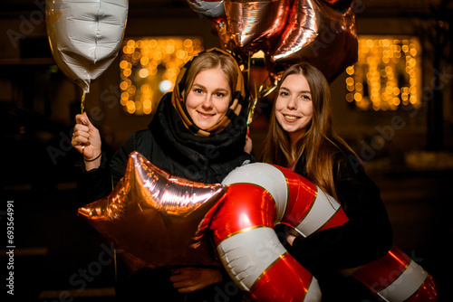 Two smiling women holding star and lollipop balloons against a blurred background of glowing lights