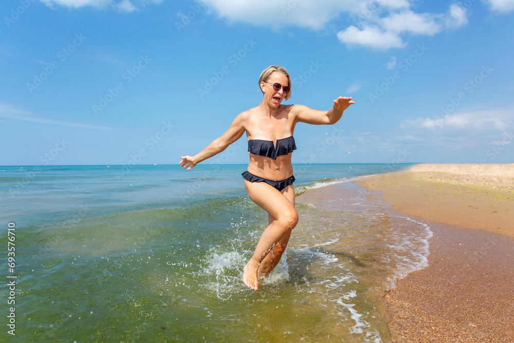 A young woman in a black bikini falls while running out of the sea on a sunny day. Activity and humor. Travel and holidays at the resort. A young woman in a black bikini falls while running out of the sea on a sunny day. Activity and humor. Travel and holidays at the resort.