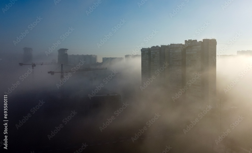 Construction cranes and a multi-storey residential building, shrouded in fog, in the early spring morning.