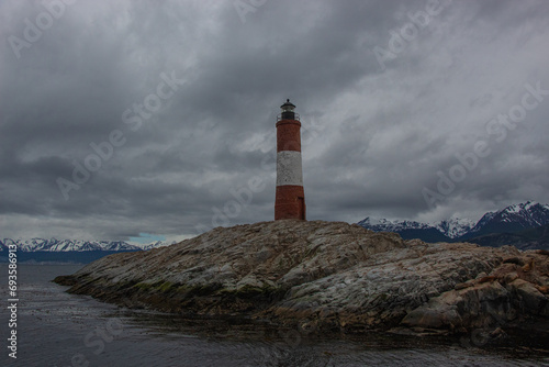 Red and white lighthouse on a small rocky island with mountain range in the background on a cloudy day