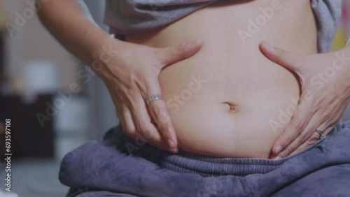 Close-up shot of her hand touching and shaking her fat belly and cellulite of an obese Asian womanin bed. The stretched skin on her abdomen emphasizes her weight gain and concerns about her health.  photo