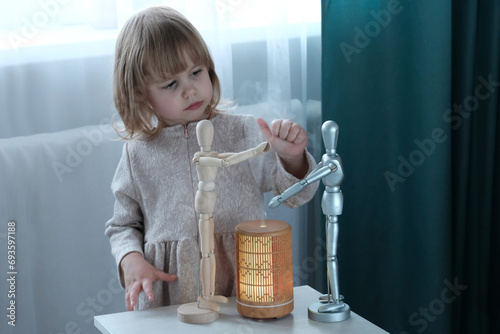 Wooden diffuser, yellow light, electric aromatic. A little blonde girl next to a wooden mannequin. The concept of care and comfort, air humidification in the house.
