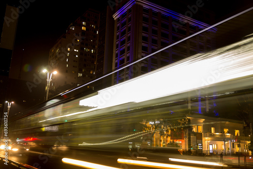 Rastro das luzes de um ônibus coletivo circulando na Avenida Paulista a noite e a fundo prédio decorado com iluminação colorida. 