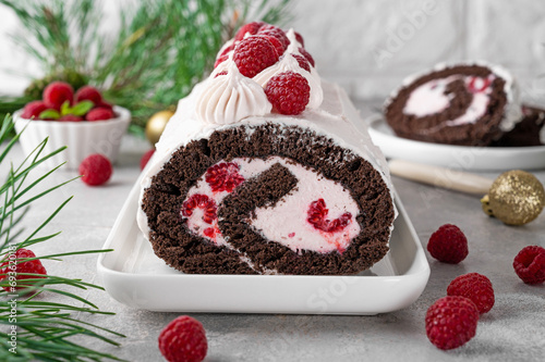 Chocolate christmas roll cake with cream and fresh raspberries on a white plate on a gray background. Christmas yule log cake.