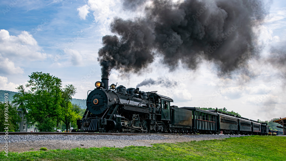 Obraz premium A View of a Narrow Gauge Restored Steam Passenger Train Blowing Smoke, Starting To Pull Out of a Station on a Summer Day