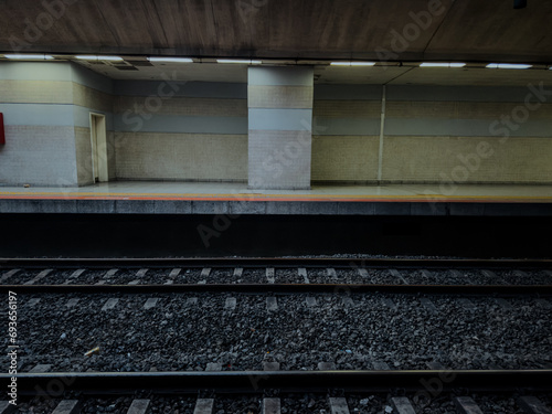 An empty, well-lit subway station platform with clean, patterned walls and two sets of tracks. The scene is desolate and quiet. railway with gravel. lonely sad concept.