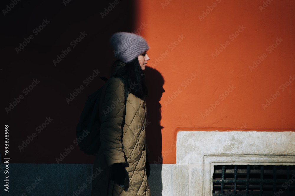 A young girl in a green down jacket and an Angora hat stands in profile near a terracotta-colored wall in the rays of the setting sun with a dark shadow. Lviv, Armenian courtyard.