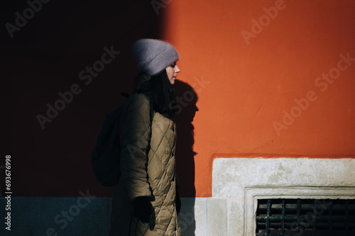 A young girl in a green down jacket and an Angora hat stands in profile near a terracotta-colored wall in the rays of the setting sun with a dark shadow. Lviv, Armenian courtyard.
