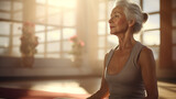 A slender modern European woman about 70 years old, well-groomed, with gray hair, sits in a calm meditative pose in a bright yoga studio filled with sunlight. Healthy lifestyle at different ages