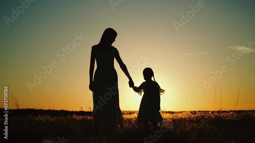 Silhouettes of mamma holding hand of daughter and talking about childhood while walking with child in sunlit field. Silhouettes of mamma with daughter sharing secrets at setting sun in park photo