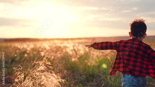 Active boy runs along field of cultivated plants to horizon in countryside at sunset. Warm sun rays fall on running boy on sunny autumn day. Delighted boy in plaid shirt runs enjoying happy childhood photo