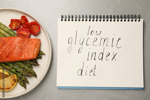 Notebook with words Low Glycemic Index Diet and plate of tasty grilled salmon on grey table, flat lay photo