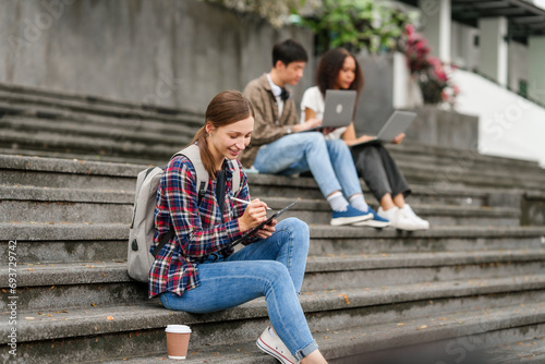 Happy diverse university students sitting on steps, using laptops and tablets, enjoying conversation. Student exchange and study abroad program, Asian man, african american woman and caucasian people.