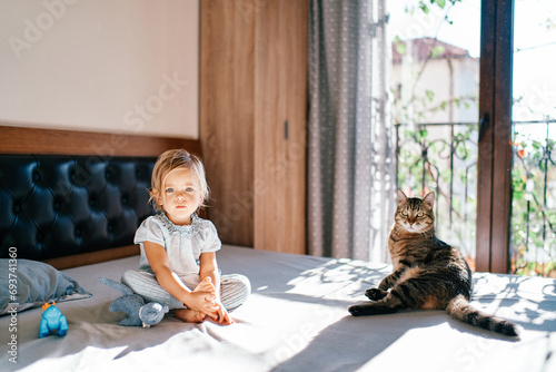 Little girl with her legs folded sits near a lying tabby cat on the bed