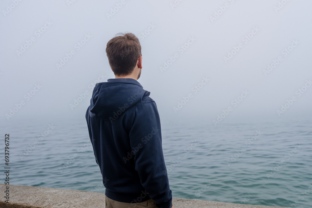 guy at sea. a young guy stands on a bridge near the sea with fog, rear view, against the backdrop of a large dark sea with white fog guy at sea. a young guy stands on a bridge near the sea with fog, rear view, against the backdrop of a large dark sea with white fog