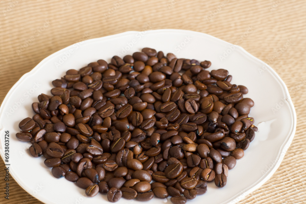 Fototapeta premium coffee beans. on a white large plate lies a large number of small brown coffee beans, the plate stands on the table, top view