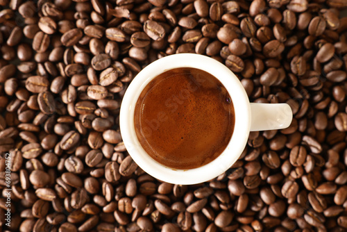 A view looking directly down on a white espresso cup filled with a freshly brewed coffee shot with brown crema. Background of roasted coffee beans.