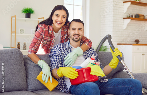 Portrait of happy smiling married couple with household cleaning supplies in hands sitting on sofa in the living room at home and looking at camera. Family housework and housekeeping concept. photo