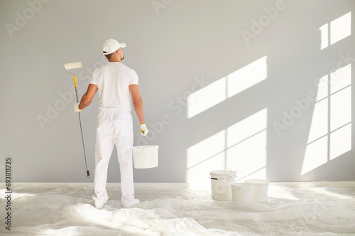 Fototapeta Naklejka Na Ścianę i Meble -  Back view of a male painter in white clothes holding a bucket and paint roller going painting a grey wall in empty room with copy space. Renovation, apartment repair and renovating house concept.