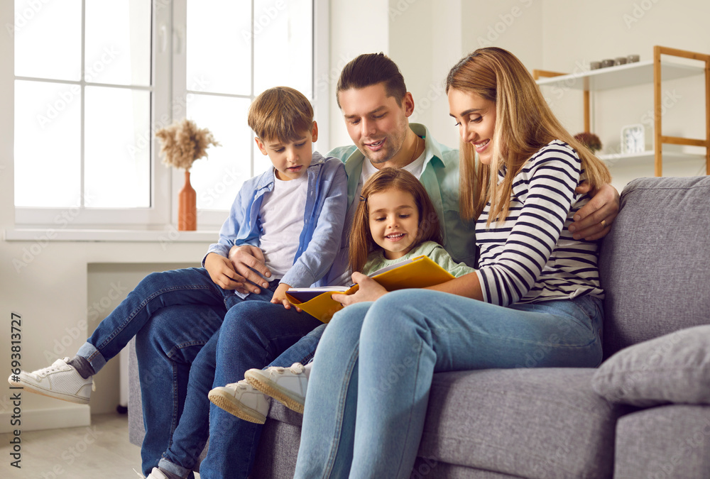 Smiling, happy family with two children are engaged in educational activities. Loving young parents with their children read book together, sitting at home on cozy sofa in the living room.