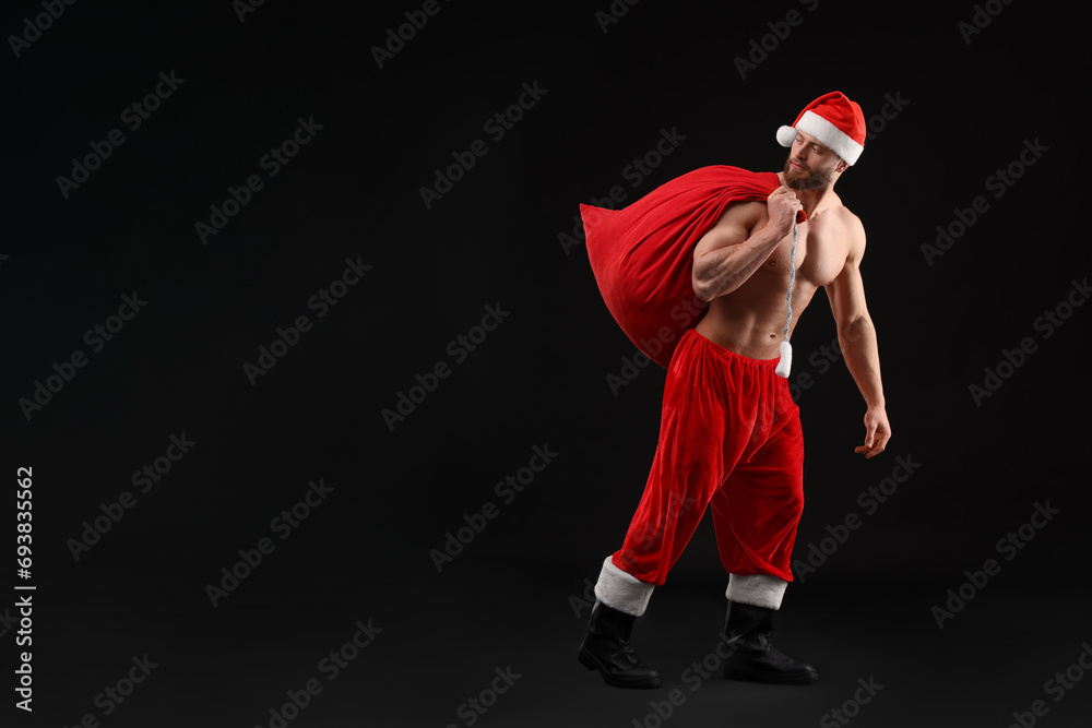 Muscular young man in Santa hat holding bag with presents on black background, space for text