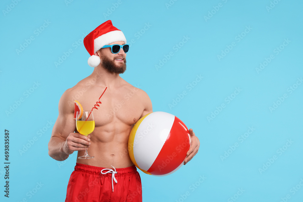 Attractive young man with muscular body in Santa hat holding ball and cocktail on light blue background, space for text Attractive young man with muscular body in Santa hat holding ball and cocktail on light blue background, space for text
