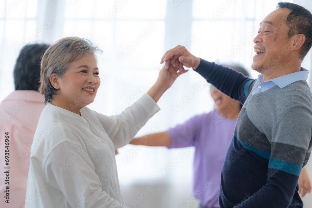 Obraz premium Asian Older male and females people dance with their partners on a dancing floor in living space. Happy older couple performing get exercise. Joyful carefree retired senior friends enjoying relaxation