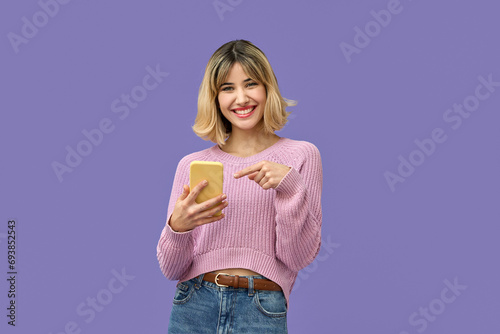 Happy pretty gen z blonde young woman model holding smartphone looking at camera pointing on cell, smiling girl using mobile apps on cell phone standing isolated on purple background with cellphone.