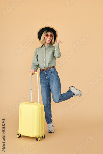 Happy pretty gen z blonde young woman, smiling cool girl tourist wearing hat and sunglasses with yellow travel suitcase waiting for vacation standing isolated on beige background. Full length.