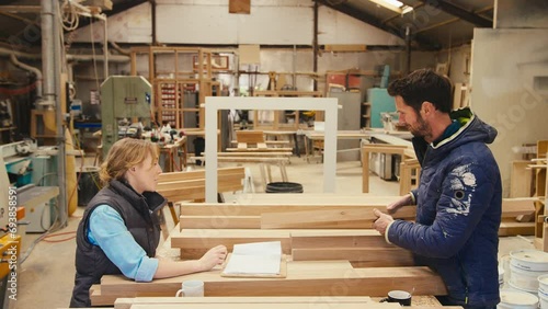 Male carpenter choosing wood and discussing drawing or plan with female trainee in woodwork woodshop - shot in slow motion photo
