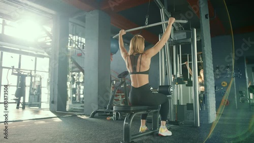 Exercise of wide grip vertical pull. A light-haired girl is working out in a gym bathed in sunlight streaming through large windows. High quality 4k footage. photo