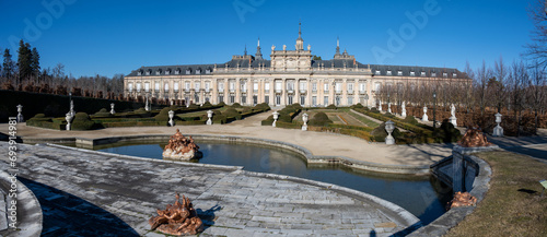 Panoramic view of the Royal Palace of La Granja of San Ildefonsoa with classical architecture and a formal garden with statues under a clear blue sky.