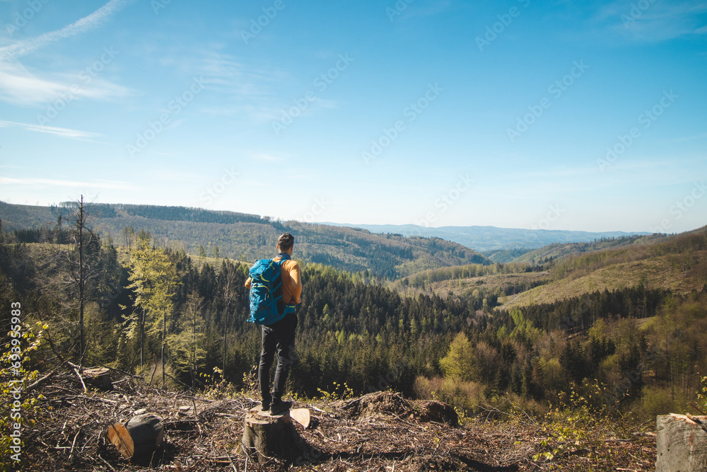 Obraz premium Adventurer and traveller with blue backpack standing on a stump in Beskydy mountains, Czech Republic. Hiking trail. Enjoying the success of magical nature
