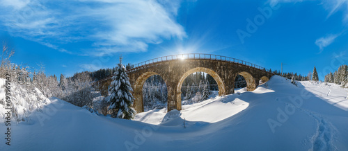 Stone viaduct (arch bridge) on railway through mountain snowy fir forest. Snow drifts on wayside and hoarfrost on trees and sunshine in sky.