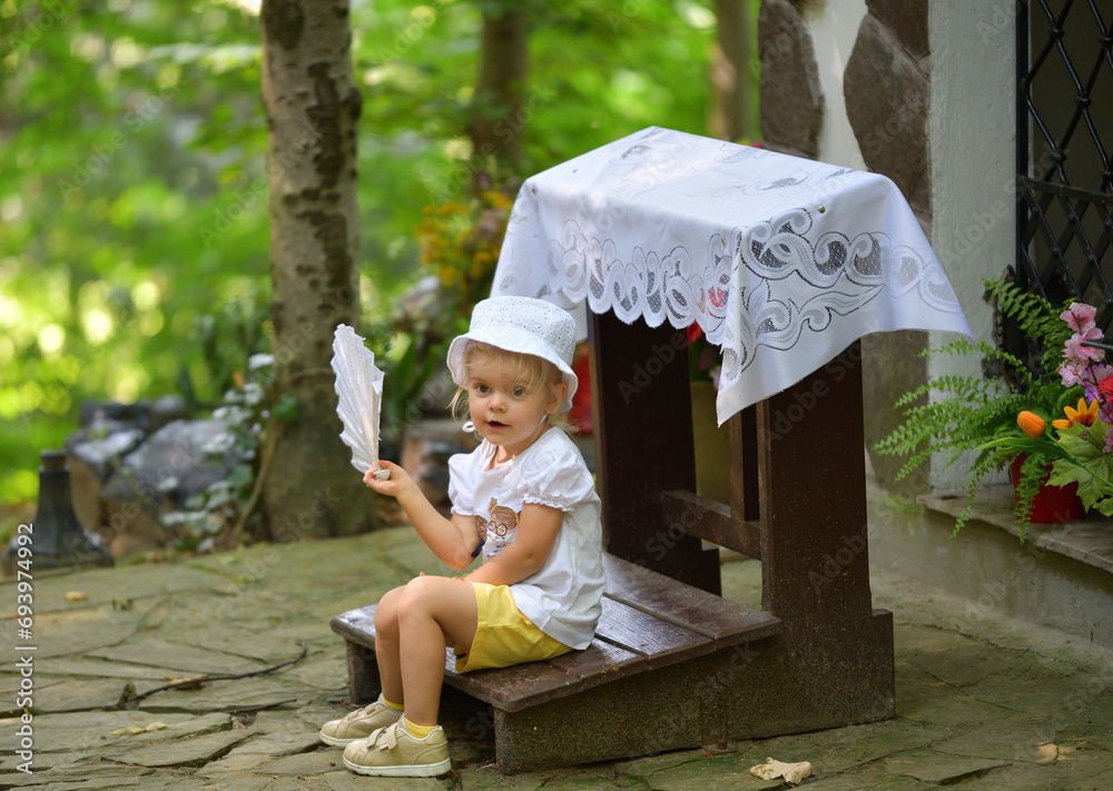 A little girl is sitting in the forest and waving a cloth fan in front of her to cool off