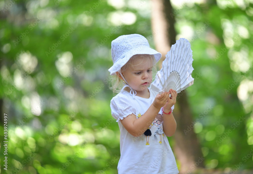 The little girl sits by the forest chapel and waves her hand with a fan like a princess