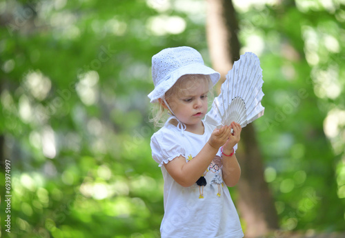 The little girl sits by the forest chapel and waves her hand with a fan like a princess