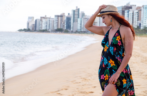 mujer con sobreso y vestido negro con flores en playa, arena y mar de fondo edificios de ciudad