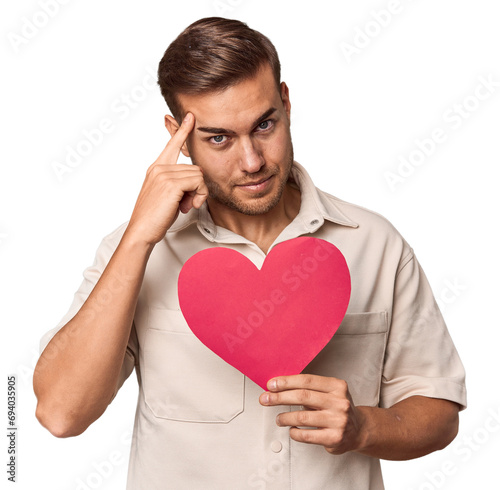 Young man holding a heart pointing temple with finger, thinking, focused on a task. © Asier