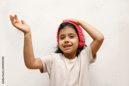 Cheerful Spanish girl child in a beige T-shirt and a pink headband on a white background, she shows with her hand, close-up, photo