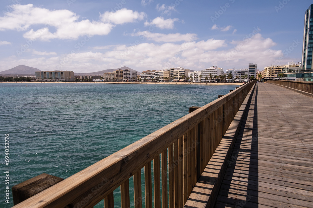 Obraz premium View of the city of Arrecife from the Fermina islet, from a wooden bridge. Turquoise blue water. Sky with big white clouds. Seascape. Lanzarote, Canary Islands, Spain.